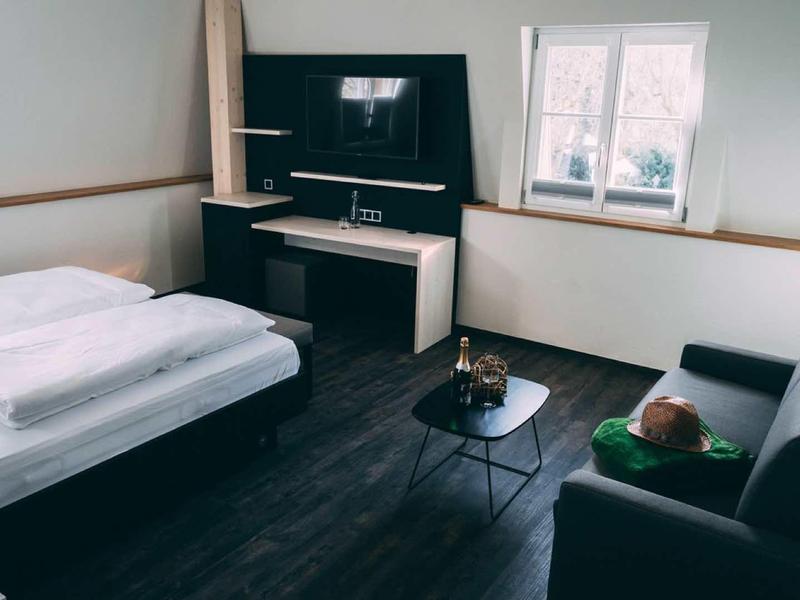 Kleines Zimmer mit Bett, Fernseher, schwarzem Sofa und rundem Tisch, helle Wände und Fenster.