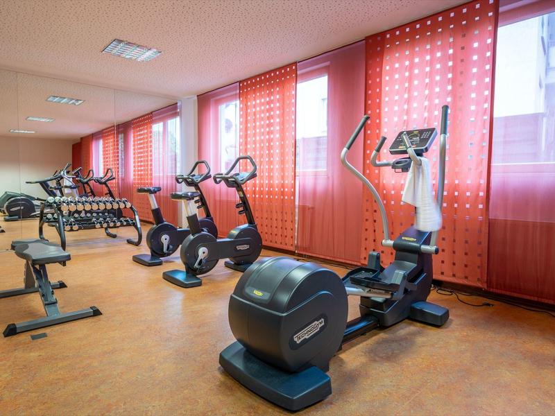 Fitnessraum mit mehreren Ergometern, roten Vorhängen und braunem Bodenbelag in hellem Raum.