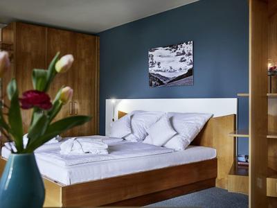Gemütliches Schlafzimmer mit Holzmöbeln, blauem Akzentwand, Blumen in Vase und weißer Bettwäsche.
