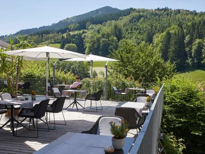 Terrasse mit weißen Sonnenschirmen, Tischen und Stühlen vor bewaldeten Hügeln unter blauem Himmel.
