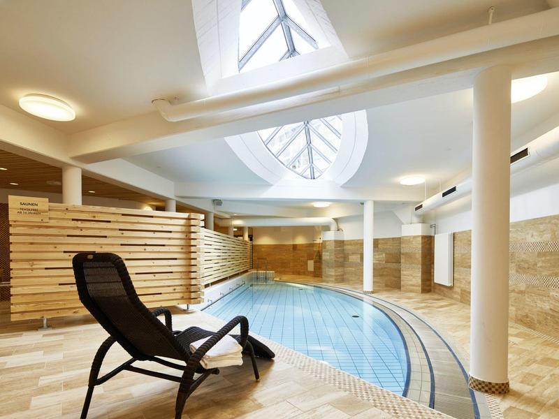 Moderner Indoor-Pool mit Holzverkleidung, beiger Steinwand und schwarzem Liegestuhl am Beckenrand.