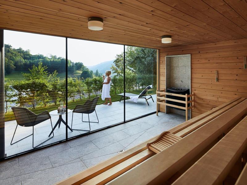 Moderne Sauna mit Holzbänken, großer Glasfront und Terrasse mit Stühlen und Blick ins Grüne.
