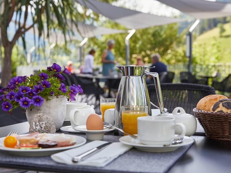 Tisch mit Frühstück, Orangensaft, Ei, Gebäck, Blumen und Tassen im Freien bei Tageslicht.