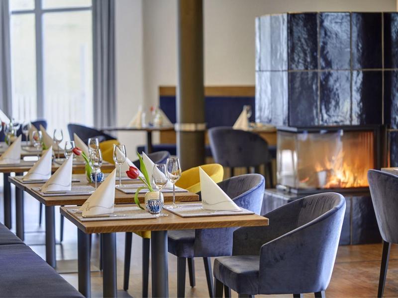 Elegantes Restaurant mit gedeckten Tischen, grauen Stühlen und einem brennenden Kamin im Hintergrund.