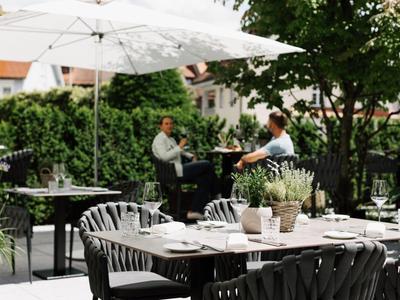 Außenbereich eines Cafés mit gedeckten Tischen, Sonnenschirm und zwei Gästen im Gespräch.