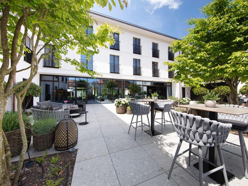 Moderner Außenbereich mit Sitzgelegenheiten, Tischen, grünen Bäumen und klarem blauem Himmel.