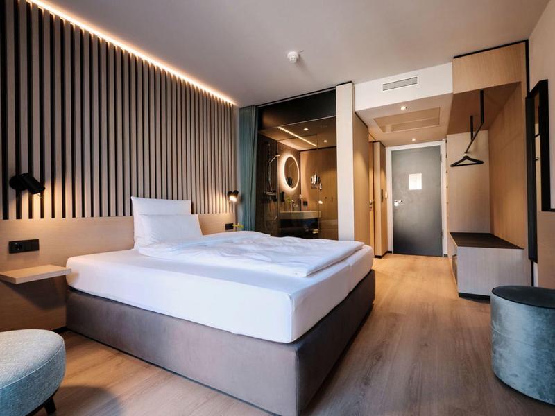 Modernes Hotelzimmer mit Doppelbett, Holzboden, indirekter Beleuchtung und Glaswand zum Badezimmer.