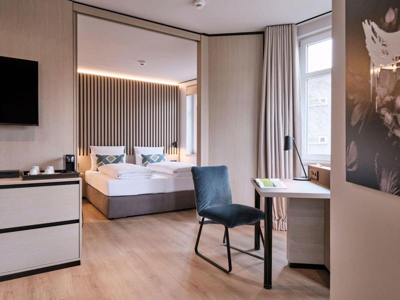 Modernes Hotelzimmer mit großem Bett, Schreibtisch, Stuhl und Fernseher an heller Wand.