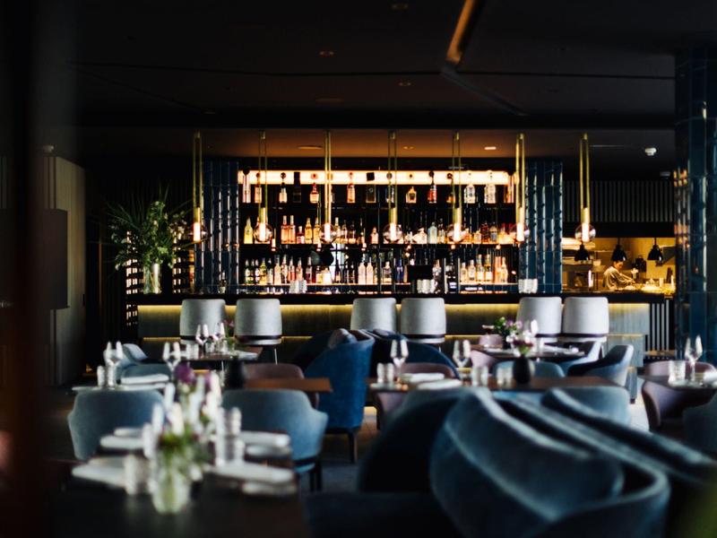 Elegantes Restaurant mit gedämpfter Beleuchtung, weißen Tischdecken und Holzdeko am Tresen.