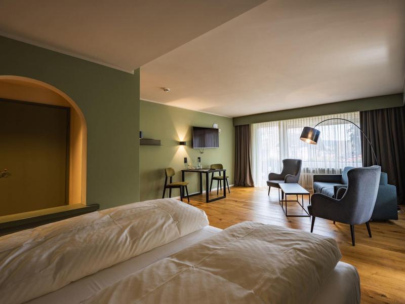 Helles Hotelzimmer mit zwei Betten, Sesseln, Fernseher und großem Fenster mit Vorhängen.