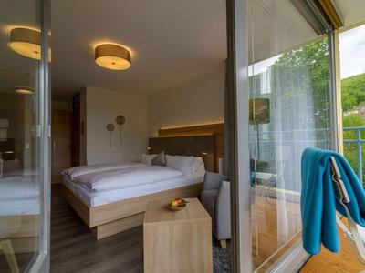 Modernes Hotelzimmer mit Doppelbett, Nachtisch und Zugang zu Balkon mit Blick auf grüne Hügel.