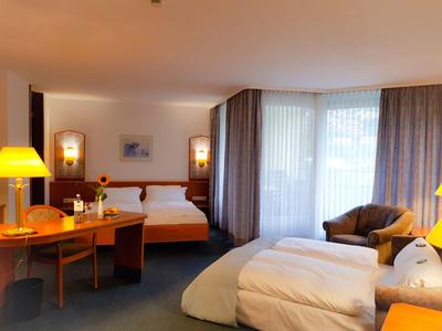 Helles Hotelzimmer mit zwei Betten, Schreibtisch, Sesseln und großen Fenstern mit Vorhängen.