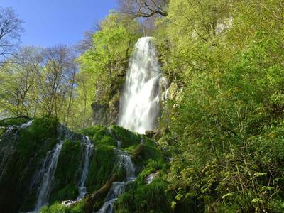 Wasserfall umgeben von grünem Laub in einem bewaldeten Gebiet unter blauem Himmel