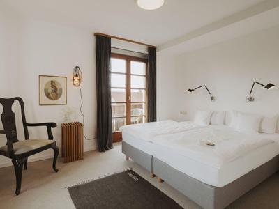 Helles Hotelzimmer mit Doppelbett, Holzstuhl, Fenster mit Vorhängen und Wandlampen.