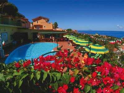 Taras hotelowy z basenem, zielonymi parasolami i czerwonymi kwiatami pod niebieskim niebem.