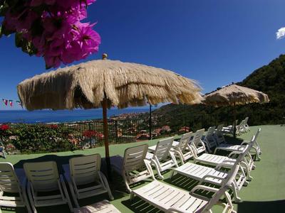 Parasolki z trzciny i leżaki na tarasie z widokiem na morze i zalesione wzgórza.