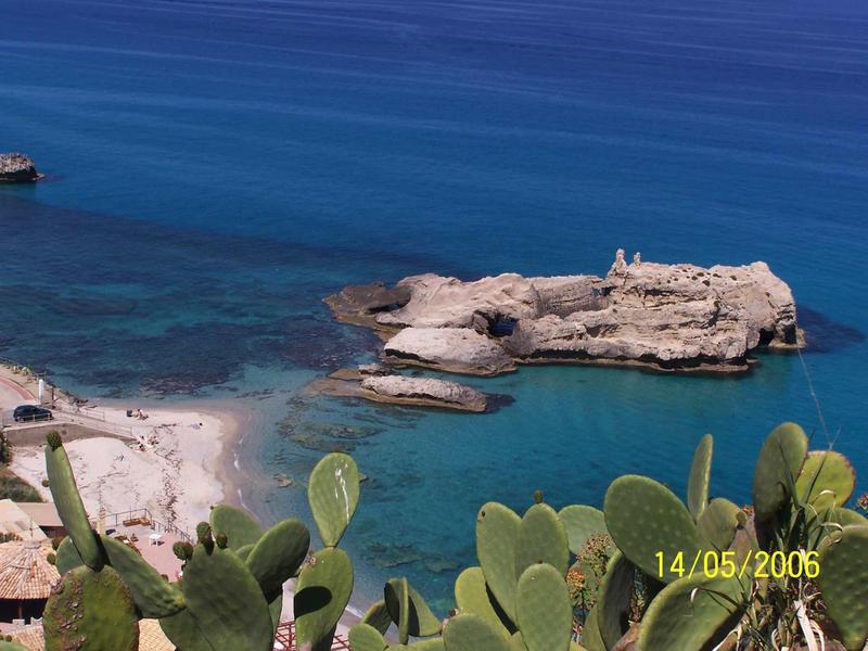 Małe skaliste wysepki i piaszczysta plaża z przezroczystą niebieską wodą i kaktusami na pierwszym planie.