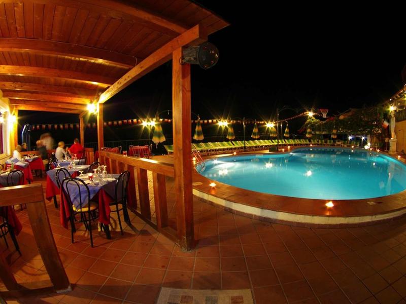 Oświetlony basen i taras hotelowy z gośćmi jedzącymi kolację na zewnątrz.