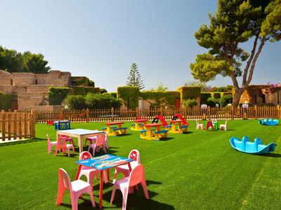 Bunter Kinderspielplatz mit Tischen, Stühlen und Schaukeln auf grünem Kunstrasen unter blauem Himmel.