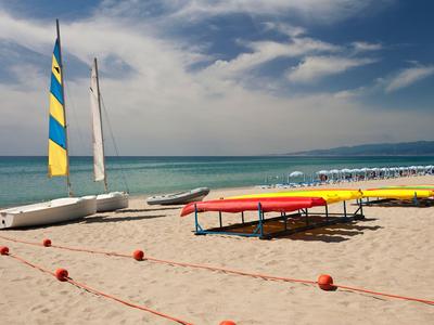 Strand mit bunten Kayaks, Segelbooten und Stühlen am klaren Meer unter einem bewölkten Himmel.
