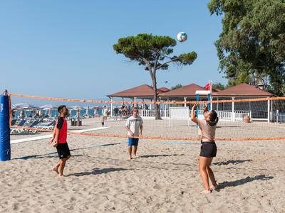 Drei Personen spielen Volleyball auf einem Sandplatz am Strand mit blauem Himmel und Hotelgebäuden im Hintergrund.