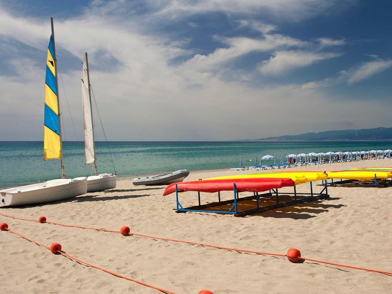 Strand mit Segelbooten, Kajaks und einer Sitzreihe am Meer bei bewölktem Himmel.