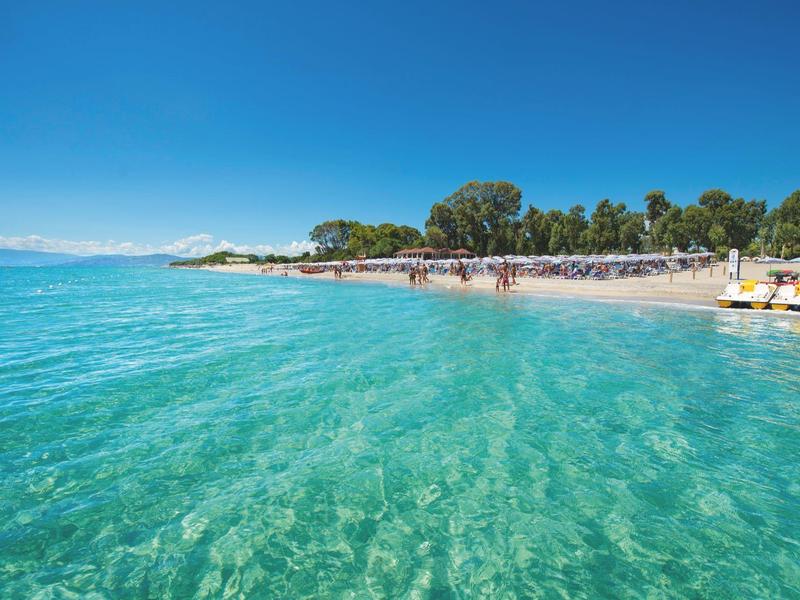 Klares türkisfarbenes Wasser am Sandstrand mit Menschen und Bäumen unter blauem Himmel.