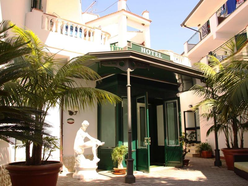 Wejście do hotelu z zielonymi drzwiami, pawilonem i rzeźbą otoczone roślinami.