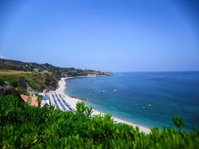 Strand mit weißem Sand, blauen Sonnenschirmen, grün bewachsenen Hügeln und klarem blauem Himmel.