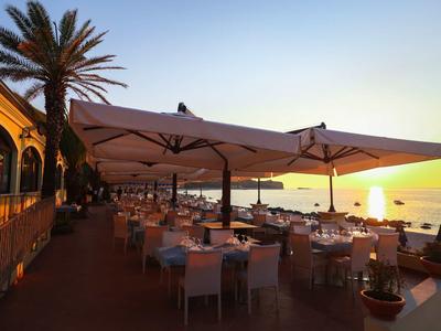 Terrassenrestaurant am Meer mit weißen Stühlen, Sonnenschirmen und Palmen bei Sonnenuntergang.