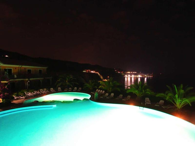 Beleuchteter Pool bei Nacht mit Pflanzen und Lichtern im Hintergrund an einem dunklen Himmel.