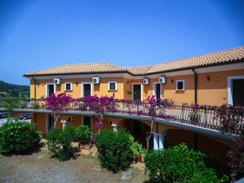 Gelbes zweistöckiges Gebäude mit dunkelrotem Dach, Balkonen und pinken Blumen unter blauem Himmel.