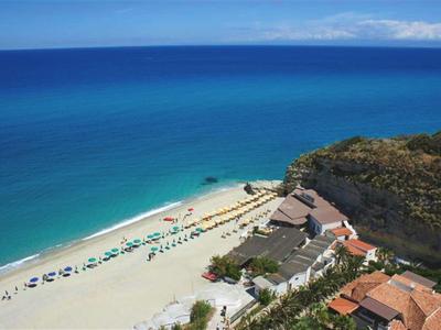 Strand mit weißem Sand, blauem Meer, Sonnenschirmen und Gebäuden neben einem steilen Felsen.