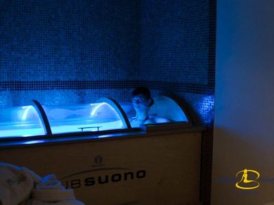 Person entspannt im beleuchteten blauen Wasser eines Holz-Wasserbeckens in dunklem Raum.