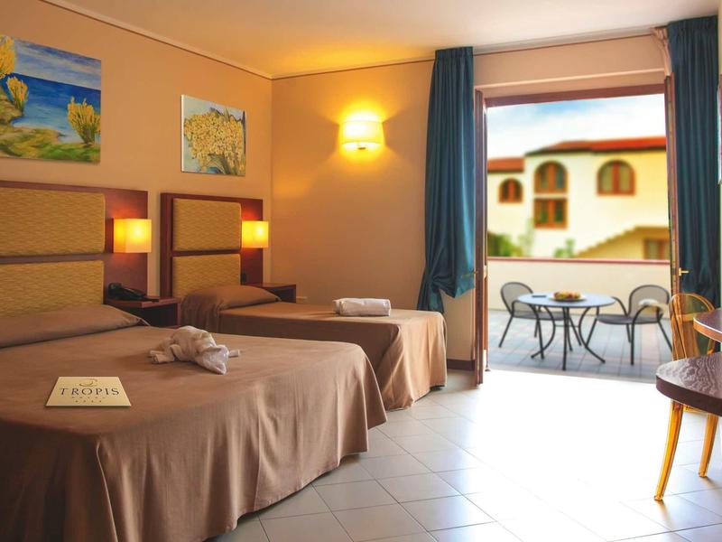 Hell eingerichtetes Hotelzimmer mit zwei Betten, Balkon und Tisch mit Stühlen.