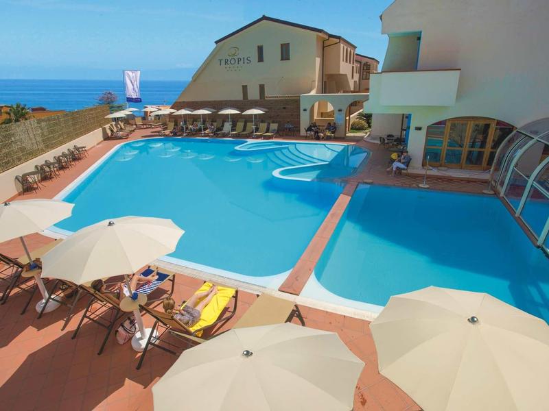 Großer Außenpool mit Liegestühlen und Sonnenschirmen neben einem weißen Gebäude mit Meerblick.