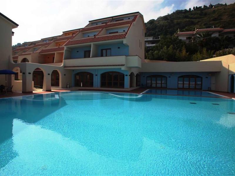 Großer blauer Swimmingpool vor einem mehrstöckigen Hotelgebäude mit Terrasse und roten Dachziegeln.
