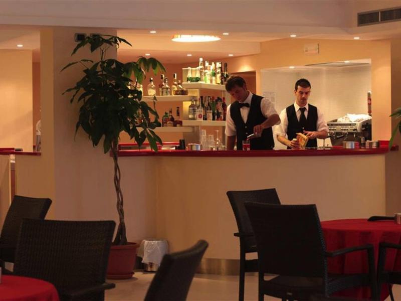 Innenraum eines Restaurants mit zwei Kellnern hinter der Theke und mehreren Tischen mit roten Tischdecken.