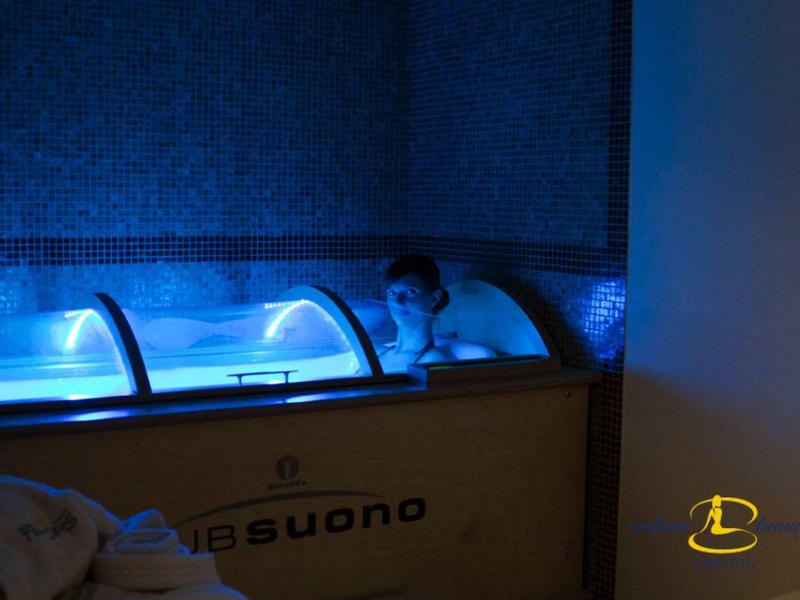 Person entspannt im beleuchteten blauen Wasser eines Holz-Wasserbeckens in dunklem Raum.