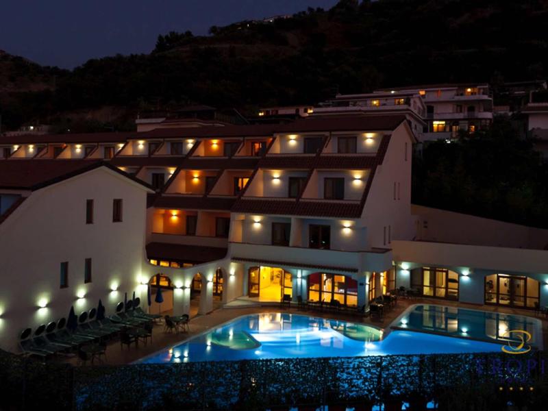 Hotel mit beleuchtetem Außenpool und mehrstöckigen Gebäuden vor dunklem Hügel in der Nacht.