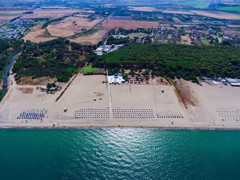 Aerial view of a wide sandy beach with sunbeds, forest and farmland behind.