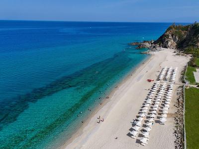 Spiaggia di sabbia bianca con file di ombrelloni accanto al mare azzurro e pendio vegetato.