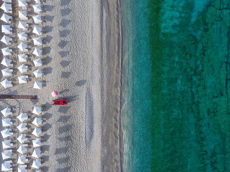 Vista aerea di una spiaggia con ombrelloni bianchi e una persona con abiti rossi.