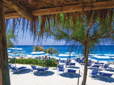Vue depuis une terrasse ombragée sur une plage avec des parasols bleus et la mer en arrière-plan.