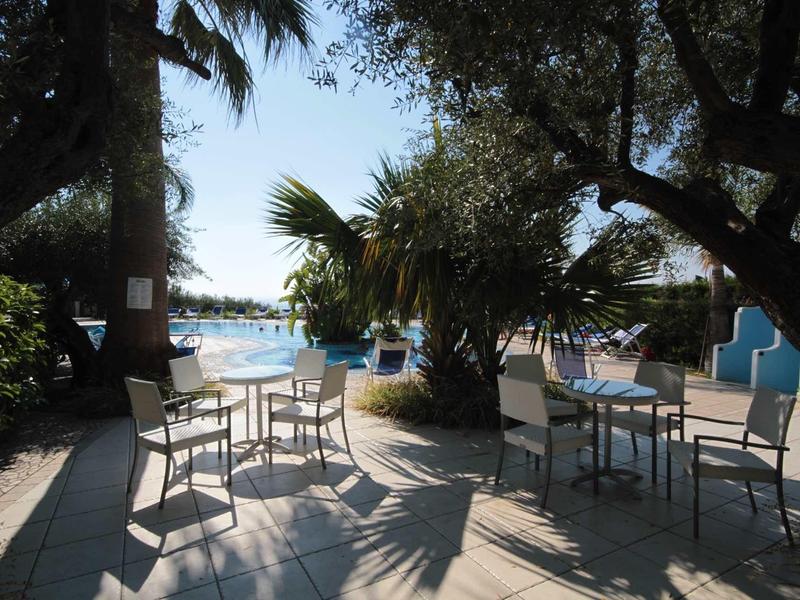 Terrasse extérieure avec chaises et tables sous les arbres près de l'eau par une journée ensoleillée.