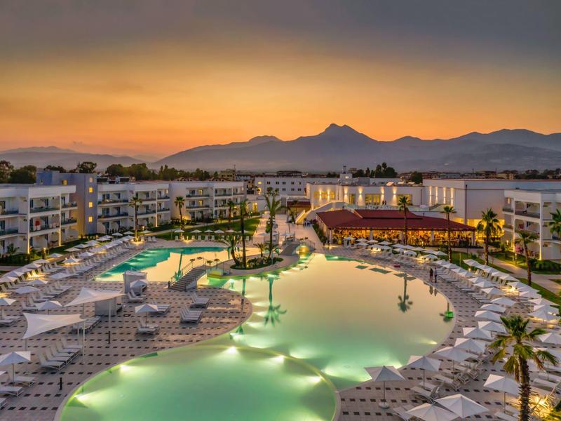 Hotel di lusso con piscina illuminata e lettini al tramonto con sfondo montano.