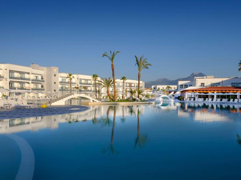 Grande piscina d'hotel con palme, lettini e edifici moderni sotto un cielo azzurro limpido.