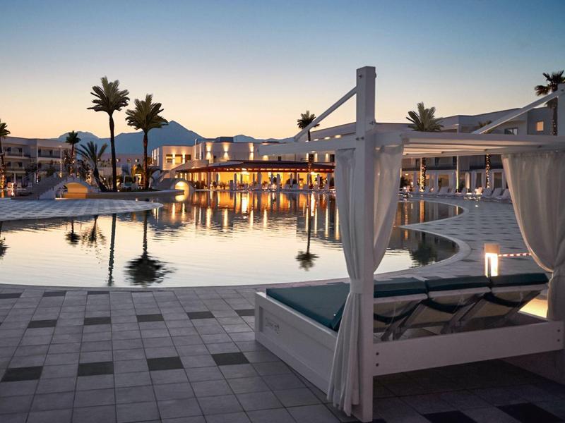 Lussuosa area piscina con letto a baldacchino e palme al tramonto in un resort.
