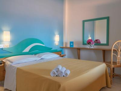 Schlafzimmer in Hotel mit Doppelbett, gelber Bettdecke, grünem Kopfteil und Spiegel mit Stuhl.