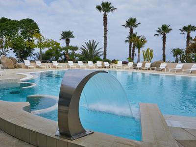 Runder Hotelpool mit Wasserfontäne, Liegestühlen und Palmen bei bewölktem Himmel.